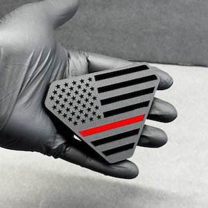May include: A gray, diamond-shaped emblem featuring a stylized American flag design. The flag includes black stripes, a field of stars, and a vibrant red line. The emblem is held in a black gloved hand.