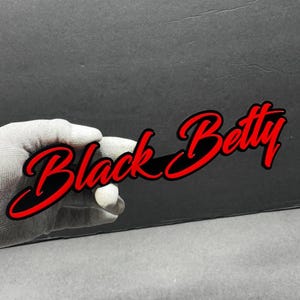 Puede incluir: Gráfico rojo y negro con las palabras "Black Betty" en fuente cursiva. El texto tiene un contorno negro y lo sostiene una mano enguantada sobre un fondo oscuro.
