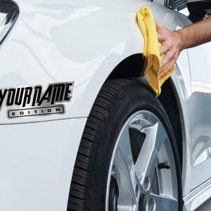 Puede incluir: Un coche blanco con una calcoman&iacute;a negra y plateada que dice "YOUR NAME EDITION".