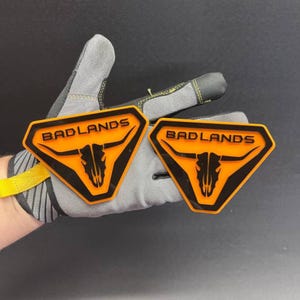 Puede incluir: Dos parches trapezoidales con un gráfico de cráneo de toro negro y el texto BADLANDS en letras negras sobre un fondo naranja brillante. Los parches tienen un borde negro grueso y un diseño moldeado en relieve con bordes geométricos definidos.