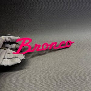 May include: A bright pink metal sign with the word "Bronco" in a cursive font. The sign is held by a gloved hand against a dark gray background. The sign is a vibrant, eye-catching piece.
