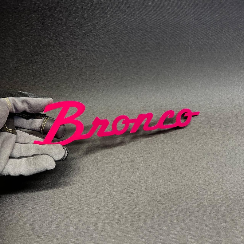 May include: A bright pink metal sign with the word "Bronco" in a cursive font. The sign is held by a gloved hand against a dark gray background. The sign is a vibrant, eye-catching piece.