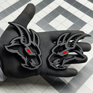May include: Two black and gray goat head emblems with red eyes. The emblems are layered with a glossy finish. The goat heads are a stylized design, likely for automotive or sports-related applications.