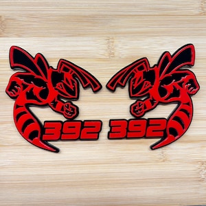 May include: Two red and black stylized hornet emblems with the number "392" in red. The emblems are on a light brown wooden surface. The hornet design features wings and a stinger.