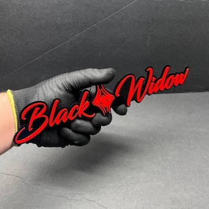 Könnte beinhalten: Ein schwarzes und rotes Schild mit den Worten "Black Widow" in einer stilisierten Schriftart. Das Schild wird von einer behandschuhten Hand gehalten. Der Text ist rot mit schwarzer Umrandung, und ein rotes Sternenmuster befindet sich zwischen den Wörtern.