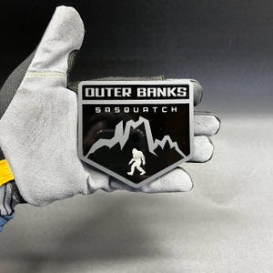 Peut inclure: Un emblème en forme de bouclier avec un fond noir, une silhouette de montagne blanche et une figure de Sasquatch blanche. La partie supérieure affiche le texte OUTER BANKS en blanc, tandis que le mot SASQUATCH est inscrit en dessous en blanc.