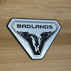 Puede incluir: Una insignia de metal plateado con un gráfico de cordillera negra y el texto "BADLANDS" en negro.