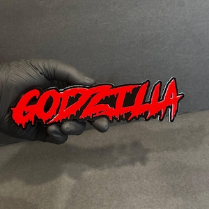 Puede incluir: Una pegatina roja y negra de "GODZILLA" con un estilo de fuente goteante. La pegatina está sujeta por una mano enguantada sobre un fondo oscuro. La palabra "GODZILLA" está en una fuente audaz y estilizada.