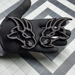 Può includere: Due emblemi a testa di capra stilizzati in bianco e nero. Gli emblemi hanno un design dettagliato con linee nitide e un contorno a contrasto. Le teste di capra sono rivolte a sinistra e sono su uno sfondo scuro.