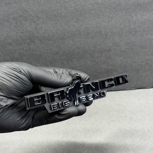 Puede incluir: Un emblema cromado con la palabra "BRONCO" y "BIG BEND" en forma rectangular. Una silueta negra de un caballo está en el centro. El emblema está sostenido por una mano enguantada.