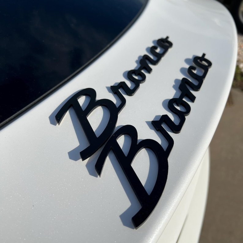 May include: Black Bronco emblem on a white vehicle. The word "Bronco" is displayed twice in a stylized font. The emblems are mounted on the rear of the vehicle, near the top.