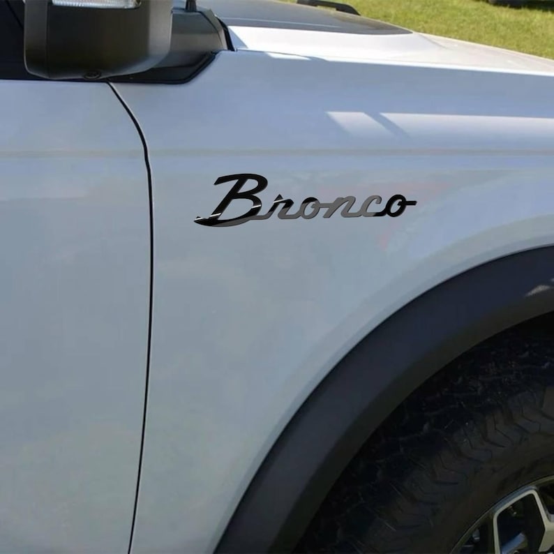 May include: Black "Bronco" emblem on a white vehicle. The emblem is in a cursive font with a shadow effect, positioned on the front quarter panel above the wheel well. The vehicle's black trim and tire are visible.