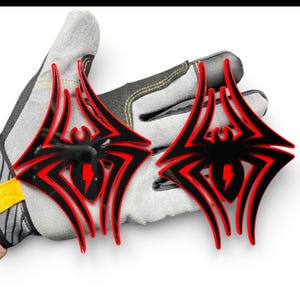 May include: Two black and red spider-shaped emblems with a lightning bolt design. The emblems are held by a gray work glove. The spider emblems have a glossy finish and are likely for automotive or decorative use.