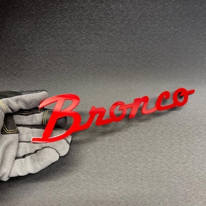 May include: A red "Bronco" emblem, with a glossy finish, is held by a gloved hand. The emblem is in a cursive font, and the letters are connected. The background is a dark gray surface.