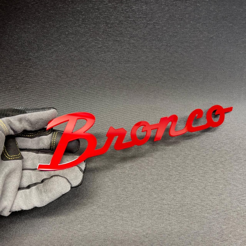 May include: A red "Bronco" emblem, with a glossy finish, is held by a gloved hand. The emblem is in a cursive font, and the letters are connected. The background is a dark gray surface.