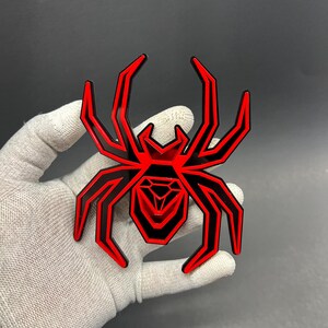 May include: A red and black spider-shaped emblem with geometric design. The emblem has a glossy finish and is likely made of metal or plastic. The spider's legs and body are outlined in black, with red filling the interior.
