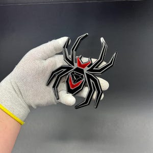Może przedstawiać: Czarny i czerwony emblemat pająka o geometrycznym wzorze. Pająk ma osiem n&oacute;g i czerwony kształt klepsydry na odwłoku. Emblemat jest trzymany w rękawicy na ciemnym tle.