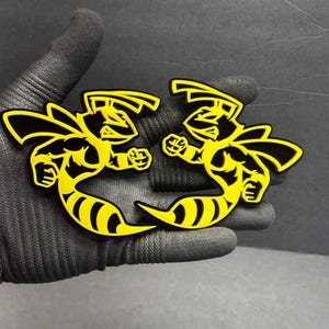 Puede incluir: Dos pegatinas con forma de abeja estilizada, amarillas y negras. Las pegatinas presentan un diseño de abeja amarilla con contornos y detalles negros. Las abejas están posadas en una posición dinámica, mirando hacia delante, con las alas extendidas y los puños apretados.