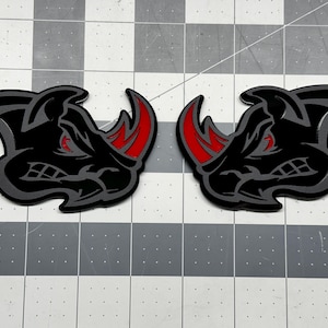 May include: Two black and grey rhino head decals with red accents on the horns and eyes. The rhino heads are facing the same direction, with a menacing expression. The decals are on a grey and white chequered surface.