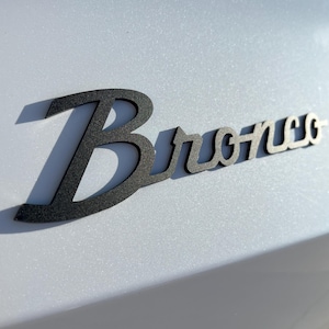 May include: A black metal "Bronco" emblem with a shadow, mounted on a light gray surface. The word "Bronco" is stylized in a cursive font. The emblem is likely for a vehicle.