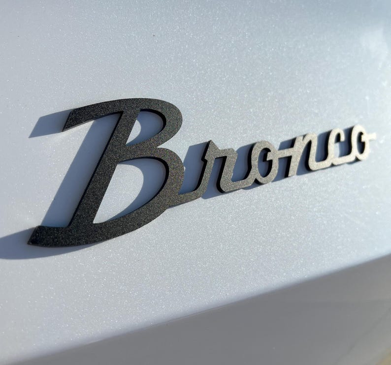 May include: A black metal "Bronco" emblem with a shadow, mounted on a light gray surface. The word "Bronco" is stylized in a cursive font. The emblem is likely for a vehicle.
