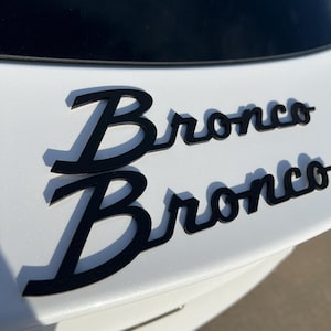 May include: Black Bronco emblem, with a shadow effect, is affixed to a white surface. The word "Bronco" is displayed twice, in a stylized font. The emblem is likely for a vehicle.