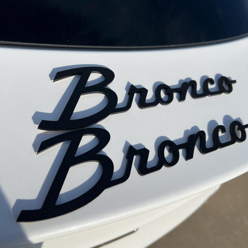 May include: Black Bronco emblem, with a shadow effect, is affixed to a white surface. The word "Bronco" is displayed twice, in a stylized font. The emblem is likely for a vehicle.