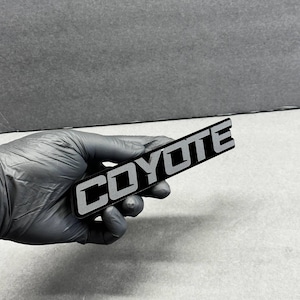 May include: A black rectangular emblem with the word "COYOTE" in silver block letters. The emblem is held by a gloved hand against a gray background. The letters have a reflective quality.