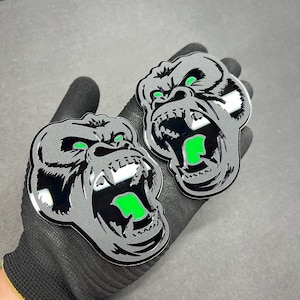 Può includere: Due adesivi argentati a forma di testa di gorilla con bocca aperta e dettagli verdi. Le teste di gorilla hanno un design dettagliato con denti affilati e sono esposte su una mano guantata di nero.