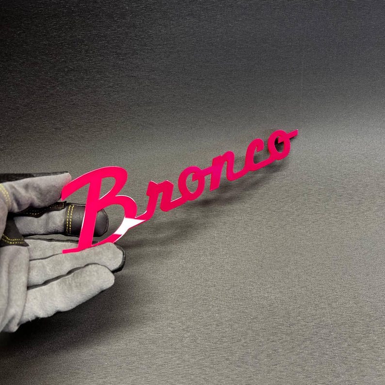 May include: A bright pink metal sign with the word "Bronco" in a cursive font. The sign is held by a gloved hand against a dark gray background. The sign is a vibrant, eye-catching accessory.