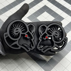 May include: Two black ram head emblems with red eye details. The emblems are held in a gloved hand against a gray and white checkered surface. The ram heads have detailed horns and a stylized design.