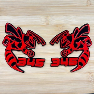 Può includere: Due emblemi rossi e neri abbinati con un design a calabrone stilizzato con il numero "345" in rosso. Gli emblemi sono posti su uno sfondo di legno marrone chiaro.