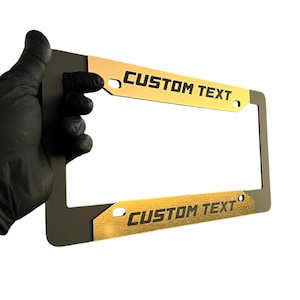May include: A license plate frame with gold-coloured accents and the words "CUSTOM TEXT" in black. The frame has a rectangular shape with a dark grey border.