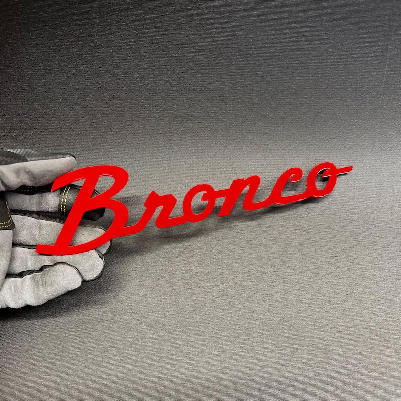 May include: A red "Bronco" emblem in a cursive font. The emblem is held by a gloved hand against a dark gray background. The emblem is a bright, solid red color.