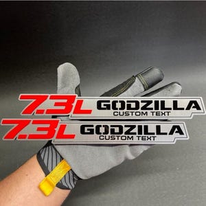 May include: Two rectangular metal emblems with the text "7.3L GODZILLA CUSTOM TEXT" in red and black. The emblems are silver with a black border. The background is dark.