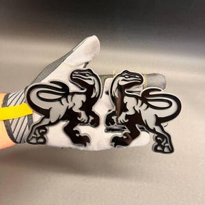 May include: Two identical dinosaur emblems featuring a glossy black silhouette layered over a matte grey base. Each emblem depicts a stylised raptor in a side profile stance with a long tail, sharp claws, and a distinct head shape, creating a layered effect.
