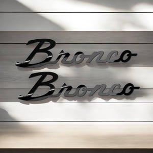 May include: Two black and gray "Bronco" wordmark emblems. The word "Bronco" is in a stylized script font. The emblems are displayed on a white and gray wooden background.