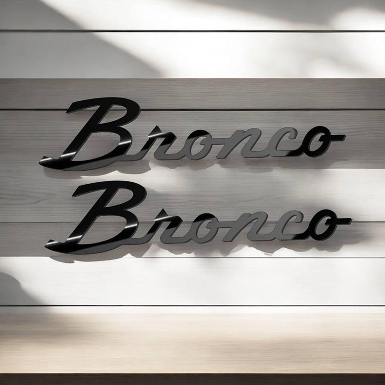 May include: Two black and gray "Bronco" wordmark emblems. The word "Bronco" is in a stylized script font. The emblems are displayed on a white and gray wooden background.