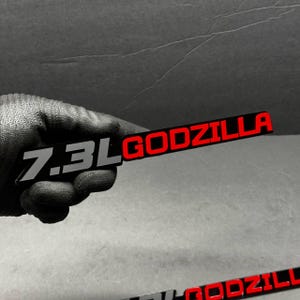 Może przedstawiać: Czarny i srebrny emblemat z napisem "7.3L GODZILLA". Słowo "GODZILLA" jest czerwone, a "7.3L" srebrne. Emblemat ma prostokątny kształt i wydaje się być wykonany z metalu.