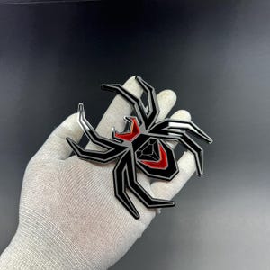 Może przedstawiać: Czarny i srebrny emblemat pająka z czerwonymi akcentami. Pająk ma geometryczny kształt z ostrymi kątami. Emblemat jest trzymany w rękawicy na ciemnym tle. Ciało pająka ma czerwony wz&oacute;r.