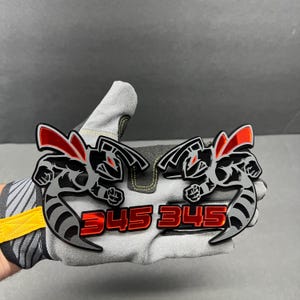 Può includere: Due emblemi automobilistici neri e rossi con un design a vespa stilizzato e la scritta "345 345" in rosso. Gli emblemi sono tenuti da un guanto da lavoro grigio.
