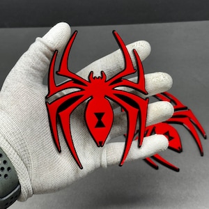 Puede incluir: Emblema de araña roja y negra con un símbolo de viuda negra en el centro. El emblema de la araña tiene patas largas y puntiagudas y se sostiene en una mano enguantada. El emblema es un diseño brillante y tridimensional.