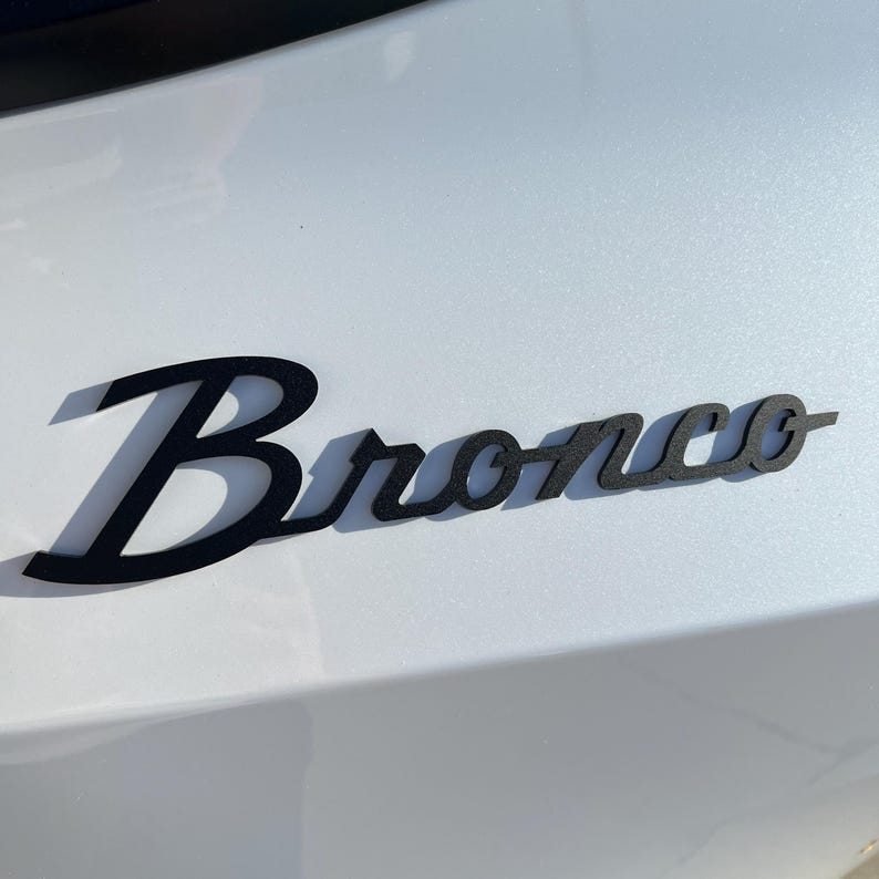 May include: Black metal "Bronco" emblem in a cursive font, mounted on a white vehicle surface. The emblem casts a shadow, enhancing its three-dimensional appearance. The image is taken in daylight.