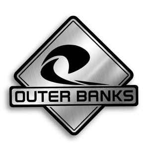 May include: A diamond-shaped metallic sign with a brushed silver finish and a black border. The center features a black graphic of a stylized ocean wave. A horizontal rectangular bar across the middle displays the text OUTER BANKS in black.