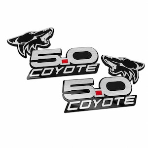 May include: Two identical automotive emblems featuring a stylized black coyote head silhouette above the text 5.0 COYOTE. The numbers 5.0 are rendered in a brushed silver finish with a small red square accent, while the word COYOTE is in white.