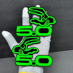 Könnte beinhalten: Zwei grüne und schwarze 5.0 Logo-Aufkleber mit einem Cartoon-Hasen mit Rennmotiv. Die Aufkleber werden von einer behandschuhten Hand vor einem dunkelgrauen Hintergrund gehalten. Der Text "5.0" ist in fetter, neongrüner Schrift.