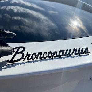 May include: A black script decal featuring the word Broncosaurus applied to the white rear exterior of a vehicle. The lettering is stylized in a cursive font and is positioned horizontally below the rear window glass of the car.