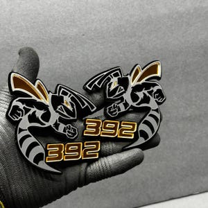 May include: Two black and silver metal emblems featuring a stylized hornet design with gold accents. The number "392" is displayed in gold lettering below each hornet. The emblems are held in a gloved hand.