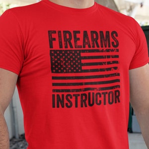 May include: Red t-shirt with a distressed American flag and the text "FIREARMS INSTRUCTOR" printed in black.