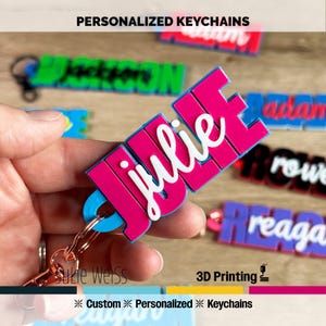 May include: A close-up of personalized keychains with names in various colors. The keychain in focus says "julie" in white script on a pink background. Other keychains with names like "jackson" and "reaga" are visible. The text "PERSONALIZED KEYCHAINS" is at the top.